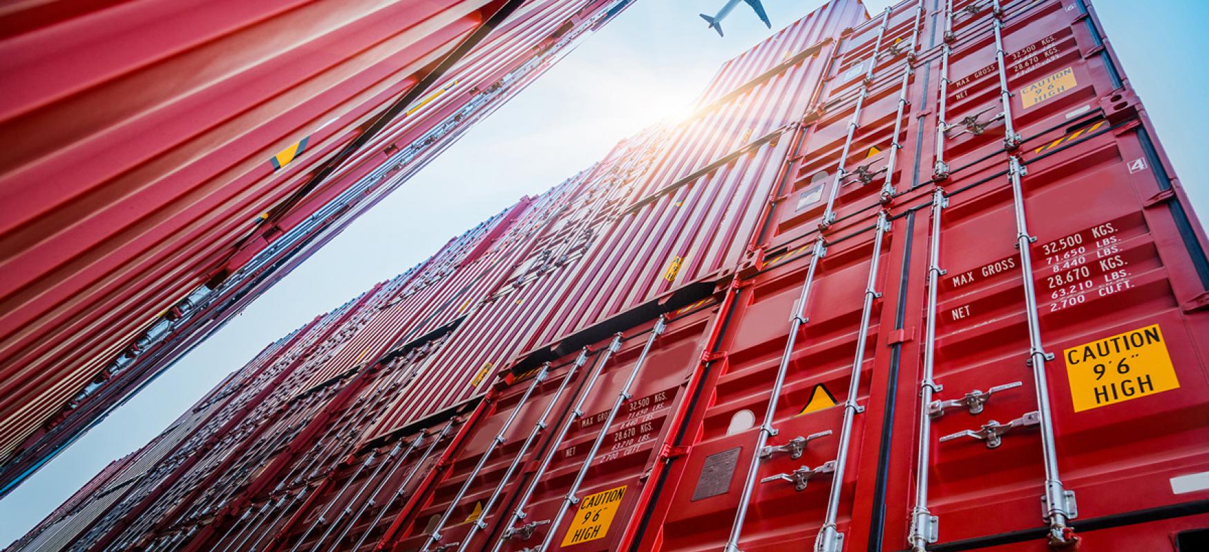 Photo of an airplane flying over stacks of red containers taken from the ground-up. 